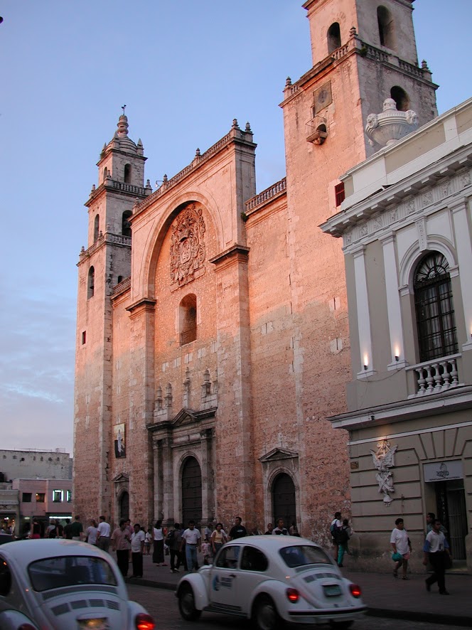 Façade of the Mérida cathedral in the evening light. Groups of pedestrians pass along the sidewalk in front as Volkswagen Beetles drive by.
