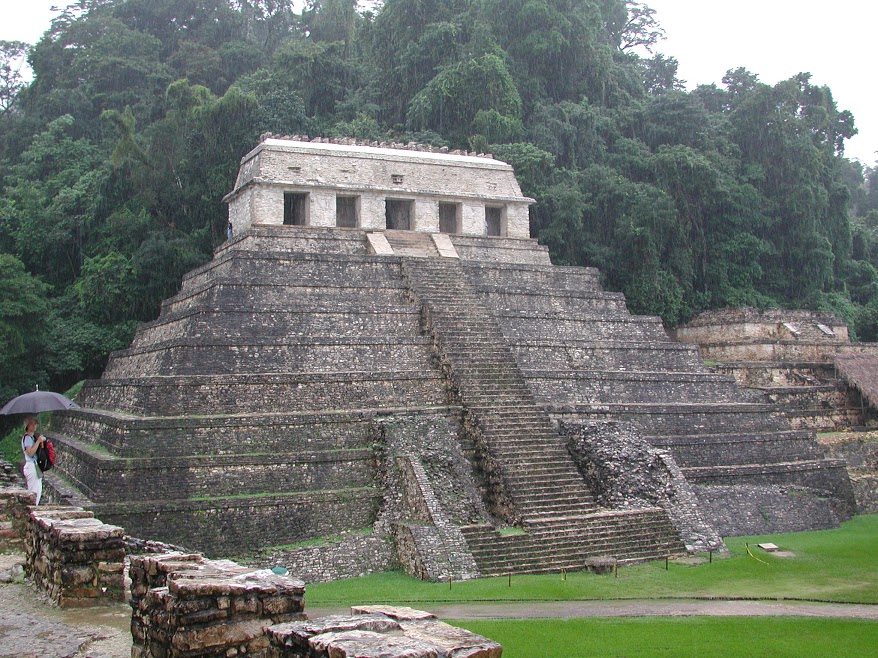 The Mayan ruins of the Temple of the Inscriptions at Palenque towering over a courtyard surrounded by jungle. A large staircase leads up the main face of the pyramid. Rain pours down in torrents.