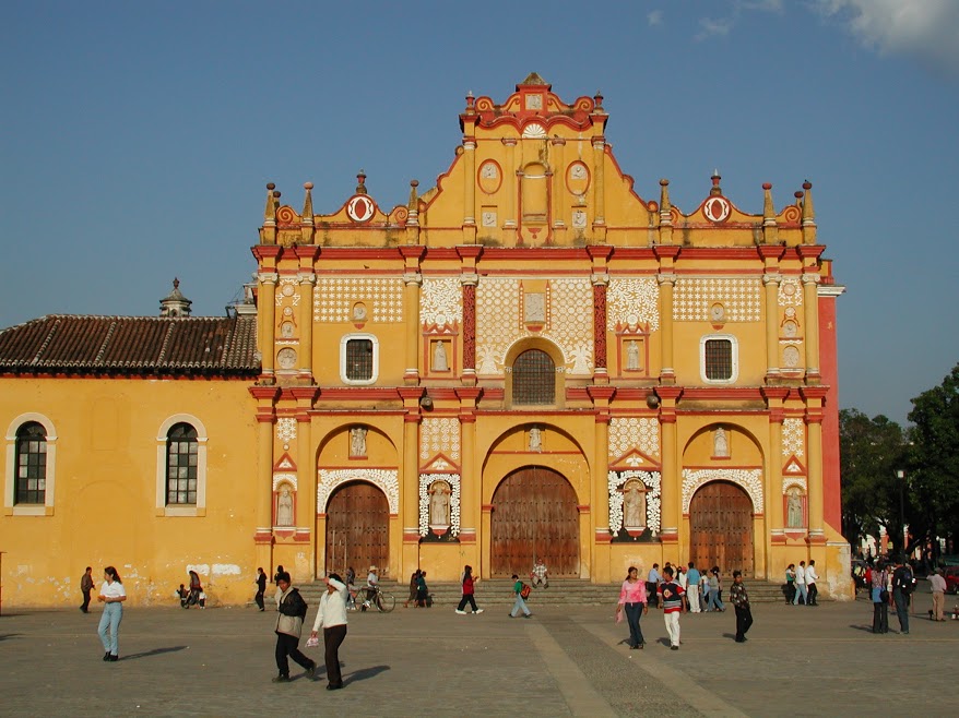 The bright yellow façade of a catheral faces the main plaza in San Cristóbal de las Casas. Pedestrials mill about the square in groups.