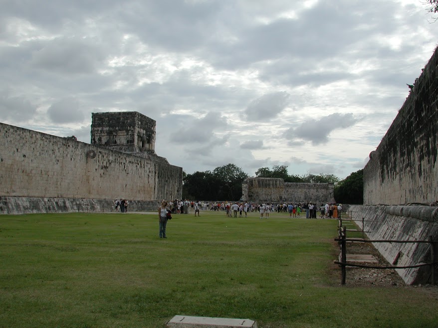 The ball court at Chichen Itzá. Large, perfectly flat stone walls rise above the grass. Two stone hoops protrude, one from each wall, facing sideways. A crowd of people stands at the far end of the court.