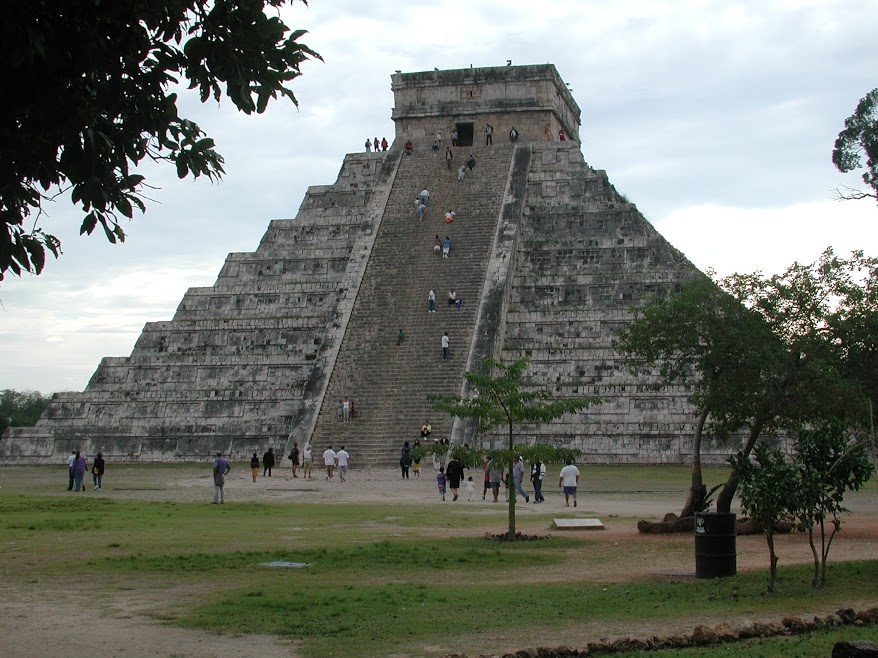 A view from the ground below the El Castillo pyramid at Chichen Itzá. Visitors climb the steep staircase leading up the centre of the face of the pyramid. A few people stand silhouetted at the top, looking down on the surrouding jungle.